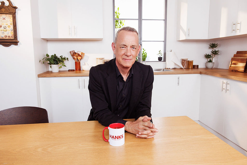 Tom Hanks sitting at a table with a 'HANKS' mug in a kitchen setting