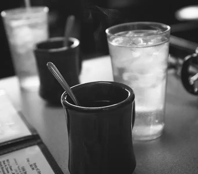 Black cup with a spoon on a table with a glass of water and other items.