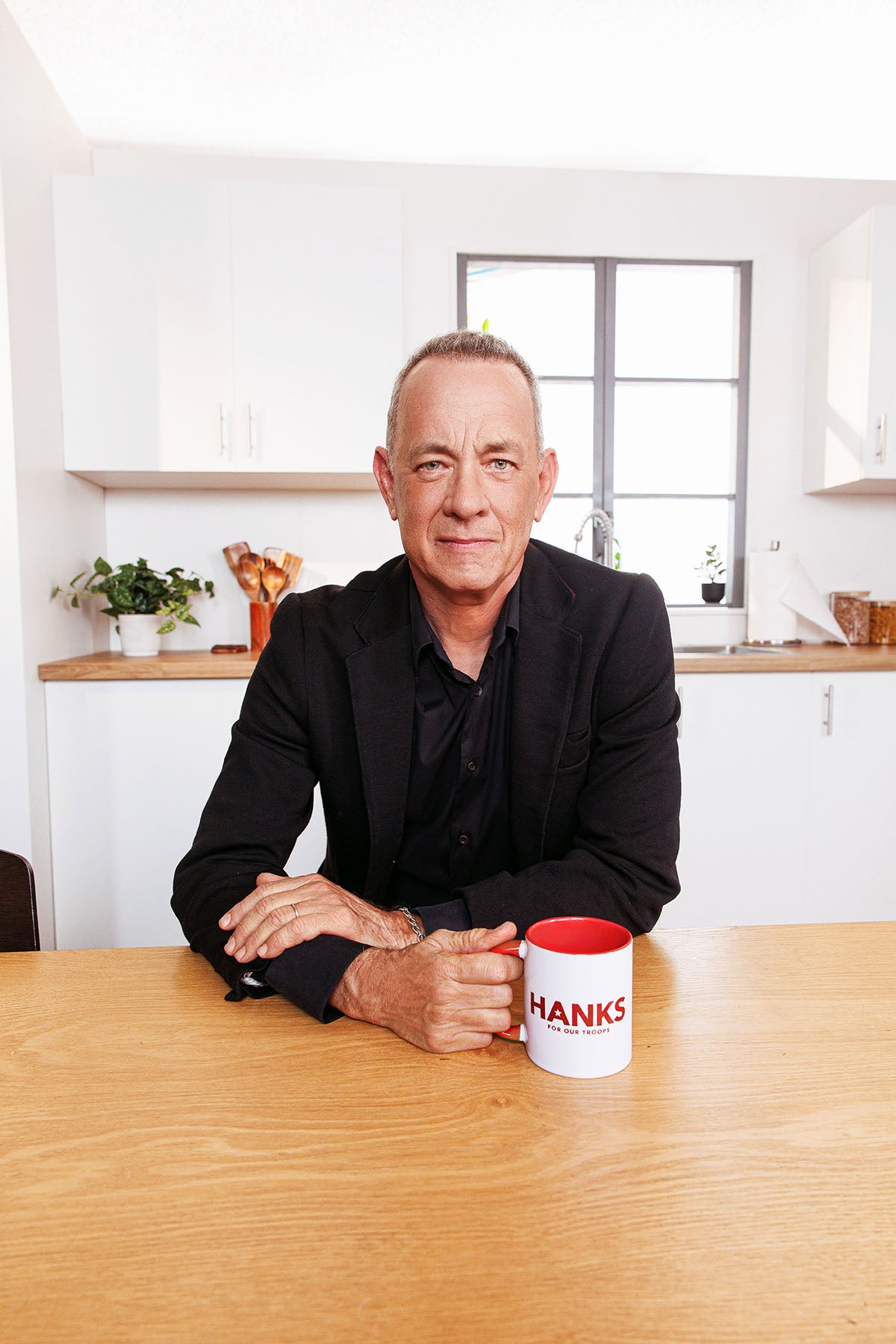 Tom Hanks sitting at a table with a mug labeled 'Hanks' in a kitchen setting